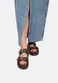Sandali neri in pelle con due fibbie regolabili, suola piatta e design aperto sulla punta. Indossati con una gonna di jeans con uno spacco laterale.
