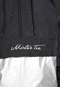 Čierny vetrovka s strieborným reflexným spodným panelom, predný zips, nastaviteľná kapucňa so šnúrkou a logo "Mister Tee" v bielej skripte.