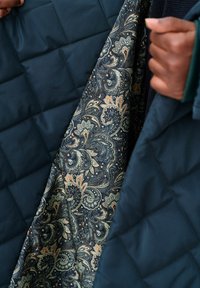 Gequilte marineblauwe buitenstof met een soepele textuur, die een binnenvoering met paisleymotief in donkerblauw, crème en oranje tinten onthult.