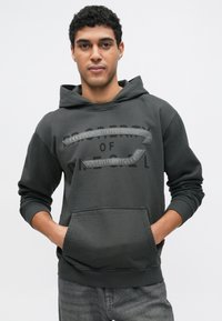 Temno siva kapa z žepom kenguruja, z reliefnimi črkami in gladkim, mehkim materialom. Nastavljiva vrvica na kapuci.