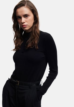 Jonge vrouw met lang golvend bruin haar, gekleed in een zwart lang mouwen top en zwarte broek met een riem, poseert met haar handen in de zakken.