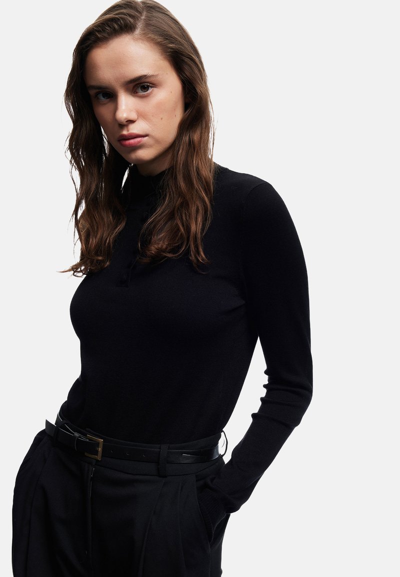 Junge Frau mit langer, welliger brauner Haar, die ein schwarzes Langarmoberteil und schwarze Hose mit Gürtel trägt, posiert mit den Händen in den Taschen.