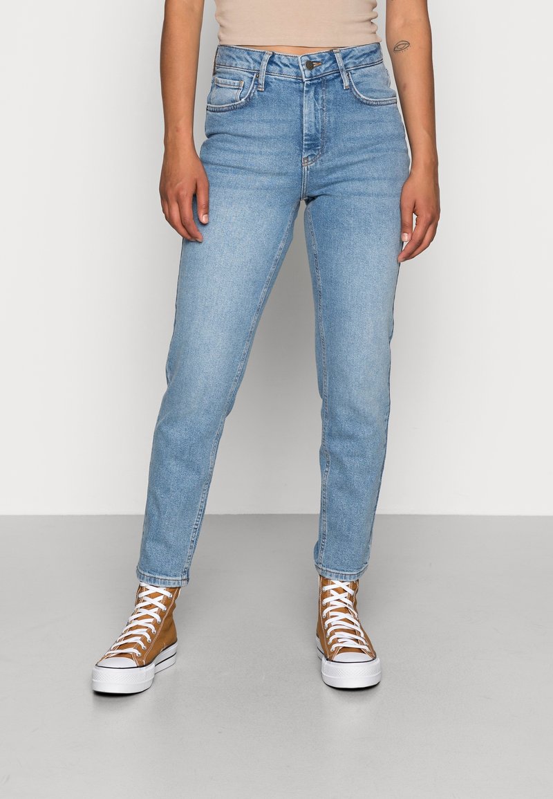 Personne portant un jean taille haute bleu clair, des baskets montantes marron à lacets blancs, debout contre un sol gris uni et un mur blanc.