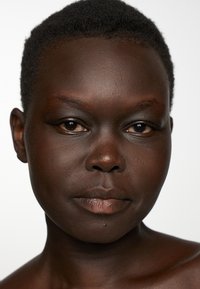 Porträtt i närbild av mörk hy med jämn struktur, kort svart hår, naturlig makeup som framhäver ögon och läppar, neutral bakgrund.