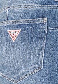 Modré džíny z denimu s kapsou na zadní straně, na které je přišitá malá trojúhelníková nášivka s logem Guess, umístěná blízko levého horního okraje.
