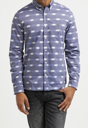 Muž nosící modrou vzorovanou košili s knoflíky na dlouhý rukáv a tmavé džíny, s jednou rukou blízko kapsy a druhou uvolněnou.