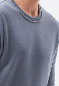 Ingrandimento di una persona che indossa un maglione a maniche lunghe, di colore azzurro chiaro e dal design semplice, su uno sfondo bianco.