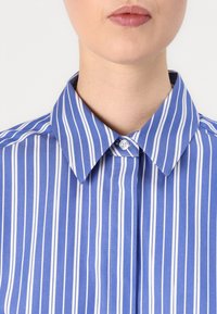Camicia a righe blu con colletto a punta, caratterizzata da sottili righe verticali bianche, chiusura con bottoni al collo e una texture del tessuto liscia.