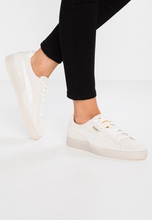 Personne portant des baskets blanches Puma Suede avec un logo doré et des leggings noirs, debout sur un sol blanc devant un fond blanc uni.