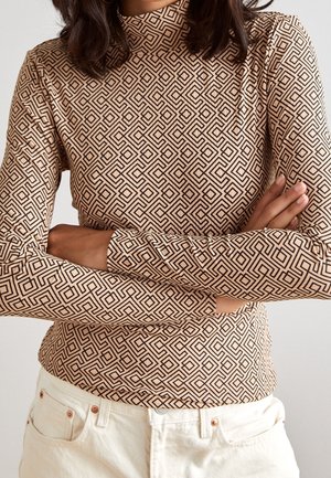 Person mit verschränkten Armen, trägt ein langärmliges beiges Oberteil mit schwarzen geometrischen Diamantmustern und cremeweiße Hose.