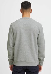 Uomo con capelli corti e ricci, che indossa una maglia a maniche lunghe grigia e semplice e pantaloni scuri, di spalle su uno sfondo bianco.