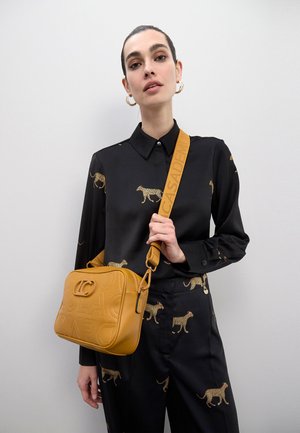 Bolso bandolera amarillo de cuero sintético con logo en relieve, correa ajustable y cierre de cremallera; llevado con una camisa negra con estampado metálico de leopardo.