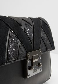 Czarna torebka z fakturowaną zamszową powierzchnią i cekinowymi akcentami, z geometrycznym wzorem oraz metalicznym zapięciem z widoczną nazwą marki.