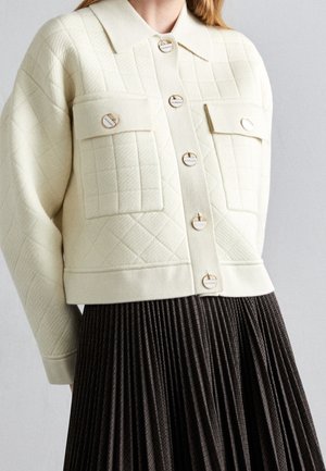 Crèmekleurig doorgestikt jack met voorflappzakken en ronde knopen, gecombineerd met een donkerbruine geplooide rok, gedragen door een persoon.