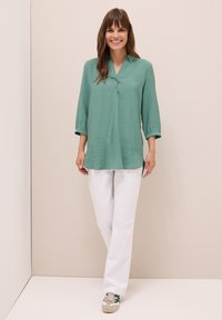 Chemisier vert à boutons avec un col en V et des manches 3/4, associé à un pantalon blanc droit et des baskets décontractées avec des accents verts.