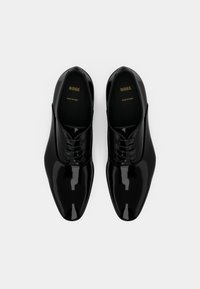 Un paio di scarpe eleganti in pelle nera lucida, con lacci e punta a punta, etichettate "BOSS Made in Italy", disposte affiancate su uno sfondo bianco.