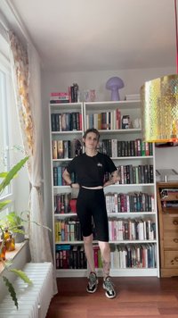 Mustat lyhyet hihatut t-paidat ja mustat pyöräilyshortsit. Harmaat lenkkarit, joissa on yksityiskohtia, tatuoituja jalkoja. Taustalla kirjahyllyjä ja ruukkukasveja.