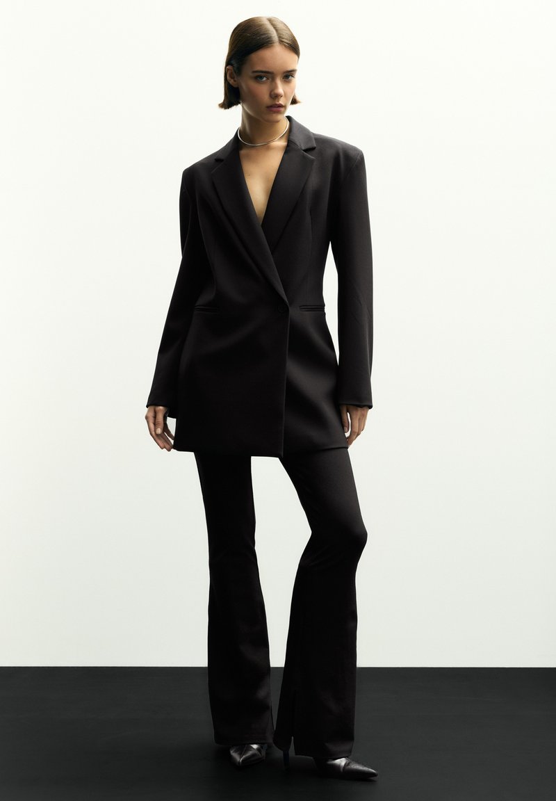 Schwarzer maßgeschneiderter Blazer mit langen Ärmeln, Reverskragen und zwei seitlichen Taschen, kombiniert mit passenden weit ausgestellten Hosen. Glatte Textur, elegantes Design.