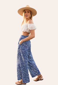 Een off-the-shoulder witte top gecombineerd met blauwe gestructureerde wijde broek; een strohoed en bruine sandalen completeren de outfit.