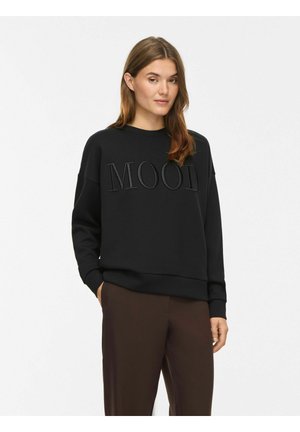 Femme aux cheveux châtain clair portant un sweat-shirt noir avec le texte "MOOI" et un pantalon marron foncé, debout devant un fond blanc uni.