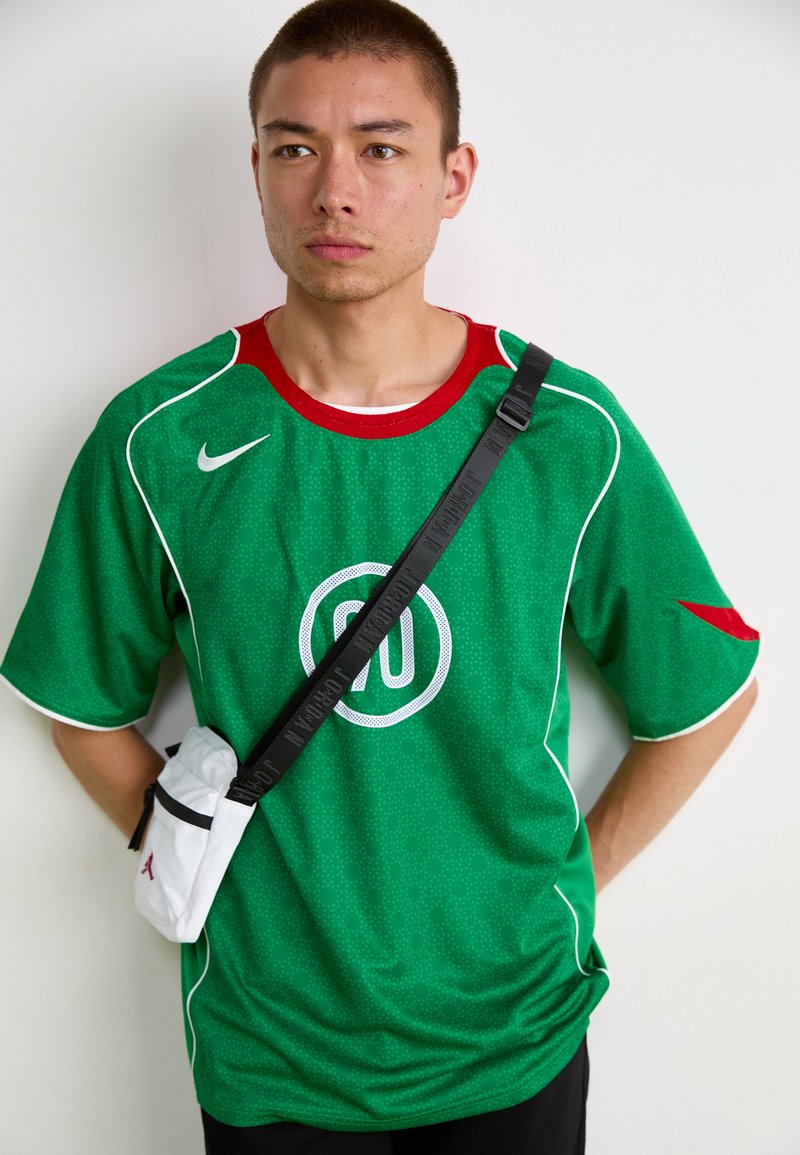 Maillot Nike vert avec col et accents rouges, doté de pipings blancs et d'un graphique circulaire. Un petit sac à bandoulière blanc ajoute une touche de détail.
