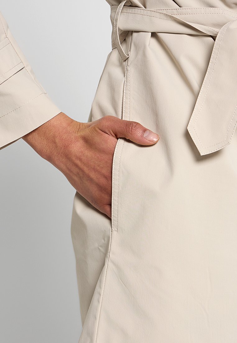 Un bolsillo de un abrigo trench beige con una mano descansando dentro. La tela parece suave, con una cintura ceñida y detalles cosidos en la costura.