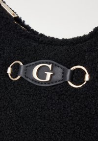 Svart teksturert stoffveske med en gullfarget bokstav "G" aksent på et svart lærreim med metallbeslag. Glidelås på toppen.