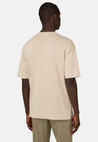 Homme portant une chemise en maille beige à manches courtes et un pantalon marron clair, vu de dos sur un fond blanc.