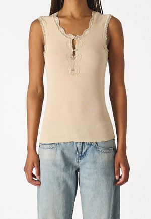 Mouwloos beige geribd topje met kantafwerking en knopen, gedragen met lichtblauwe hooggetailleerde jeans bij een staand persoon.