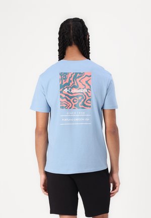 Osoba z warkoczami, ubrana w jasnoniebieską koszulkę Columbia z abstrakcyjnym różowo-niebieskim wzorem i tekstem na plecach, w połączeniu z czarnymi szortami.