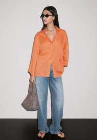 Oranje overhemd met lange mouwen, gecombineerd met een lichtblauwe brede spijkerbroek. Accessoires zijn een grijze tas en zwarte zonnebril.