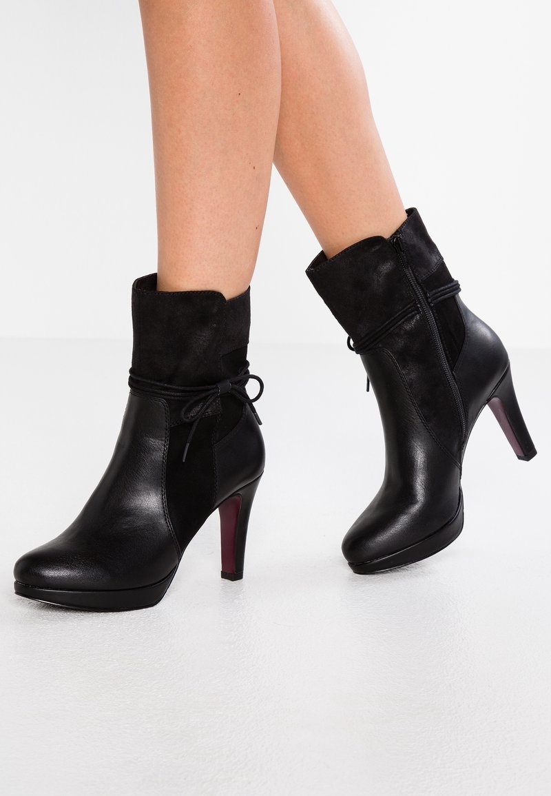 Bottes mi-mollet en cuir et suédine noire à talon avec fermeture éclair latérale et détail décoratif avec lien, portées sur jambes nues sur fond blanc.