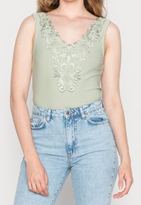 Lys grøn bodysuit med dyb V-udskæring, der har intrikate blomsterbroderier ved udskæringen; parret med lyseblå højtaljede jeans.