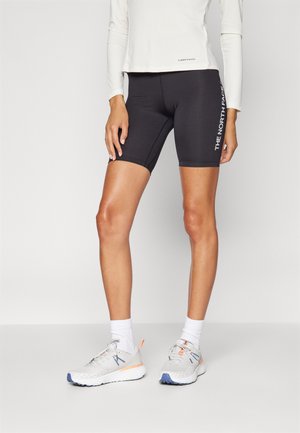 Pantaloni da ciclismo neri con dettaglio del logo sul lato, abbinati a una maglietta leggera a maniche lunghe e scarpe da ginnastica grigie con accenti colorati.