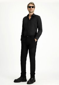 Camicia nera con bottoni e maniche lunghe, abbinata a pantaloni neri. L'outfit è completato da occhiali da sole neri e scarpe nere.