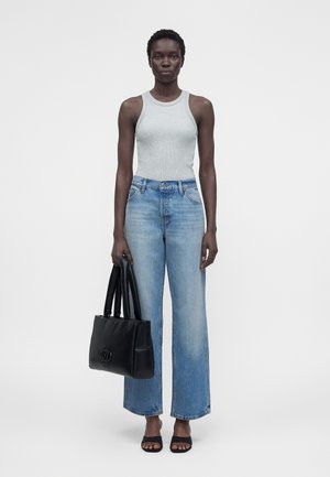 Mujer con camiseta gris sin mangas y vaqueros anchos de pierna en azul claro, sosteniendo un bolso negro y usando tacones negros abiertos en los dedos, de pie contra un fondo blanco.