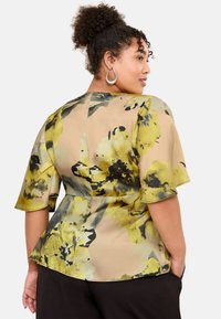 Blumenbluse mit beigem Hintergrund und großen gelben und grünen Blüten. Kurzärmelig mit tailliertem Schnitt und einem Peplum-Design.