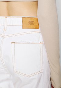 Bílé džíny z denimové tkaniny s oranžovým prošíváním, vybavené zadní kapsou a hnědou koženou nášivkou označenou "BDG." Texturovaná tkanina a výrazný tvar.