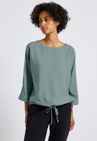 Blusa verde chiaro, dalla trama, con una vestibilità ampia, scollo largo e maniche a 3/4, dotata di un orlo con cordoncino e una leggera lucentezza.