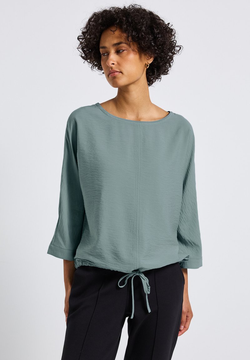 Blusa verde chiaro, dalla trama, con una vestibilità ampia, scollo largo e maniche a 3/4, dotata di un orlo con cordoncino e una leggera lucentezza.