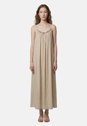 Vrouw met lang haar die een beige mouwloze maxi-jurk met dunne bandjes en sandalen draagt, staand tegen een lichte achtergrond.