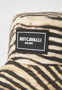 Barna kalap fekete csíkozással, fekete téglalap alakú logócímmel, fehér "JUSTCAVALLI MILANO" felirattal. Textúrált anyag.