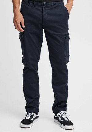 Mand iført sorte cargobukser med sidelommer og sorte sneakers med hvide snørebånd, stående mod en ensfarvet baggrund.