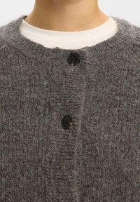 Nahaufnahme eines grauen, flauschigen Strickcardigans, der über einem weißen Rundhalsshirt am Hals und oberen Brustbereich einer Person geknöpft ist.