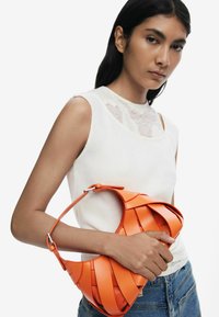 Orange woven leather bag with a curved shape. Features a short shoulder strap and a smooth finish. Model holds it against a plain background.