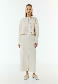 Jasna kremowa, krótka kurtka z guzikami z przodu i dwoma kieszeniami na piersi, zestawiona z dopasowaną długą spódnicą z kieszeniami bocznymi i centralnym rozcięciem.