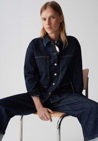 Giacca di jeans scuro con due tasche frontali, bottoni argentati e cuciture in vista. Abbinata a pantaloni in jeans scuro coordinati.