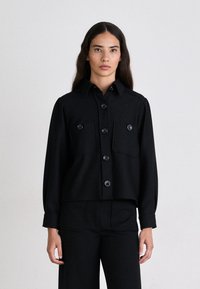 Chaqueta corta negra con cierre de botones en la parte delantera, dos bolsillos en el pecho y mangas largas. Fabricada con una tela texturizada con un ligero brillo.