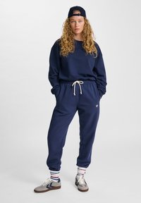 Ung person med långt lockigt hår som bär en marinblå keps bakåtvänd, marinblå sweatshirt och joggers, randiga strumpor och grå sneakers, står med händerna i fickorna.