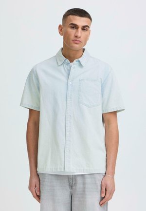 Jeune homme portant une chemise à manches courtes bleu clair boutonnée devant et un jean gris délavé, debout devant un fond uni clair.